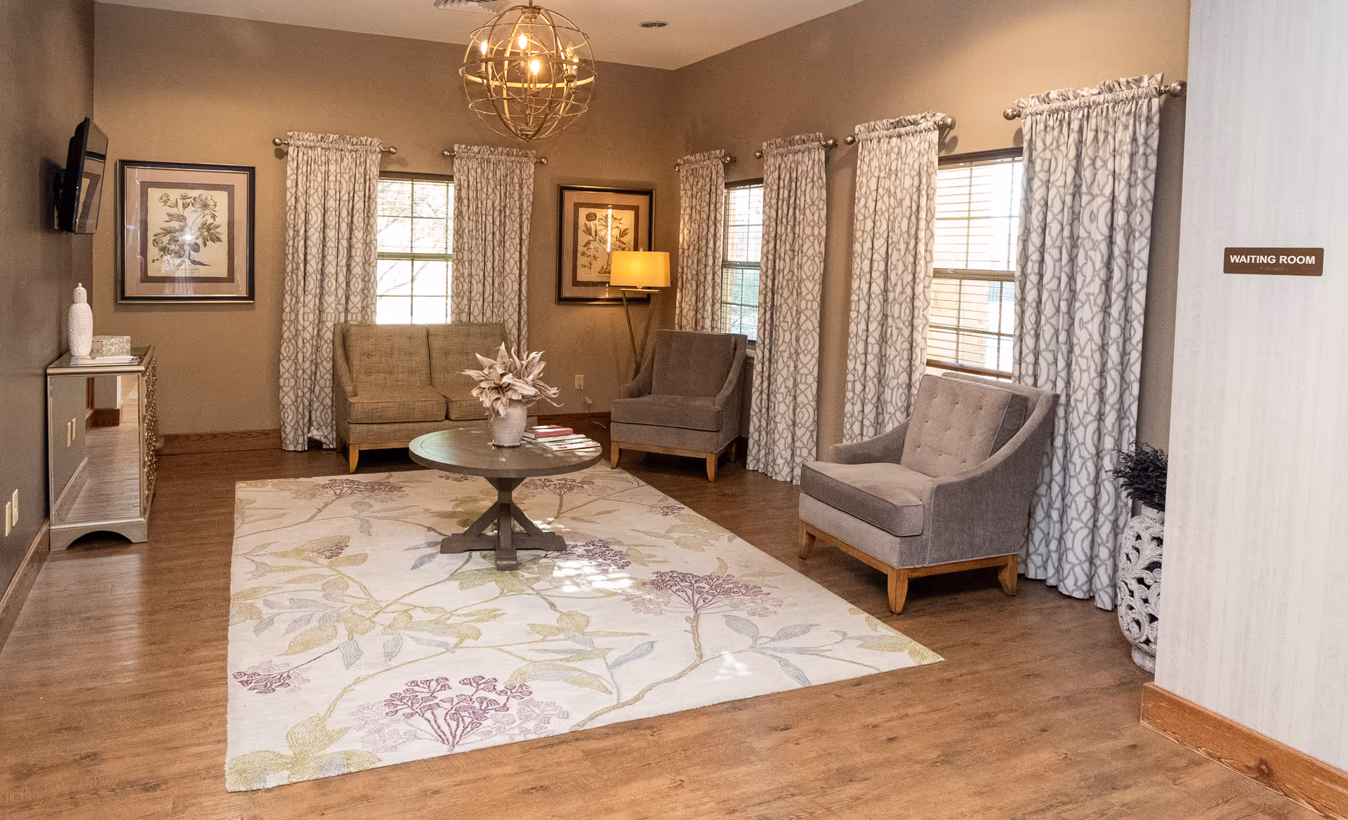 A cozy waiting room with wooden flooring, a large floral area rug, and three upholstered armchairs arranged around a round wooden coffee table with a vase of flowers. The room has multiple windows with patterned curtains, framed botanical artwork on the walls, a floor lamp, and a wall-mounted TV. A sign on the wall reads 'WAITING ROOM'.