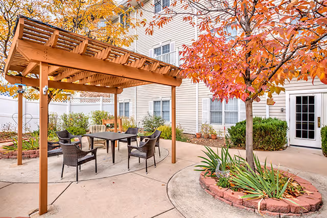 Outdoor patio area at Brookdale Monument Valley Park featuring a wooden pergola with a table and four chairs underneath. Surrounding the patio are trees with autumn-colored leaves and a multi-story building with windows and a door in the background.