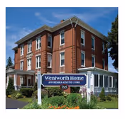 Exterior view of Wentworth Home, a three-story brick building with white trim and a covered porch, surrounded by greenery and a clear blue sky. A sign in front reads 'Wentworth Home Affordable Assisted Living 795'.