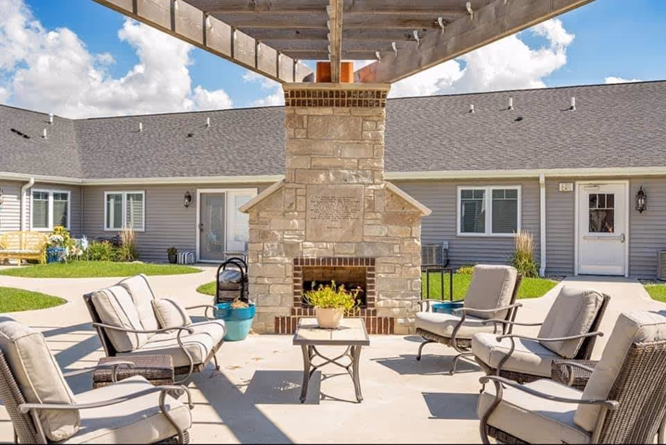 Outdoor courtyard patio with cushioned chairs arranged around a central stone fireplace in front of a single-story assisted living building.