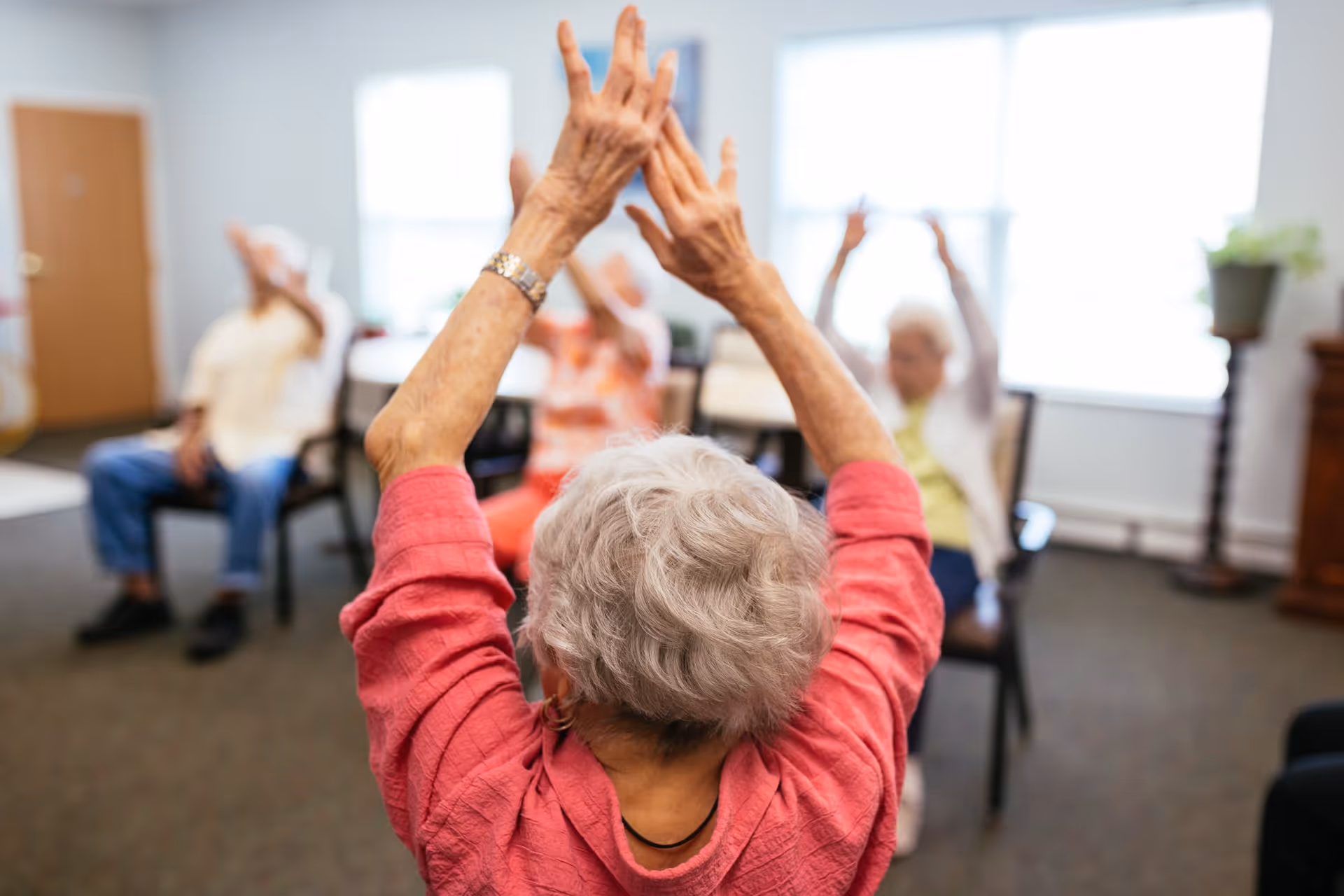 A group of elderly people sitting on chairs in a well-lit room, participating in a seated exercise or stretching activity with their arms raised above their heads.