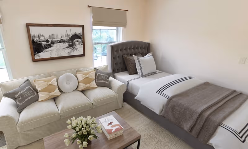 A cozy bedroom featuring a single bed with a tufted gray headboard, white bedding with black stripes, and a gray blanket. Next to the bed is a cream-colored sofa adorned with decorative pillows. A wooden coffee table in front of the sofa holds a vase of white tulips and two books. The room has light-colored walls, a window with a beige roller shade, and a framed black and white landscape photograph on the wall.