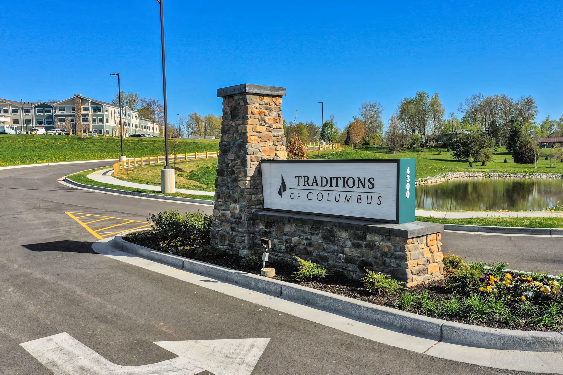 Stone and concrete sign for Traditions of Columbus at the entrance of a senior living facility, with a curved driveway, landscaped flower beds, a pond, and a multi-story building in the background under a clear blue sky.