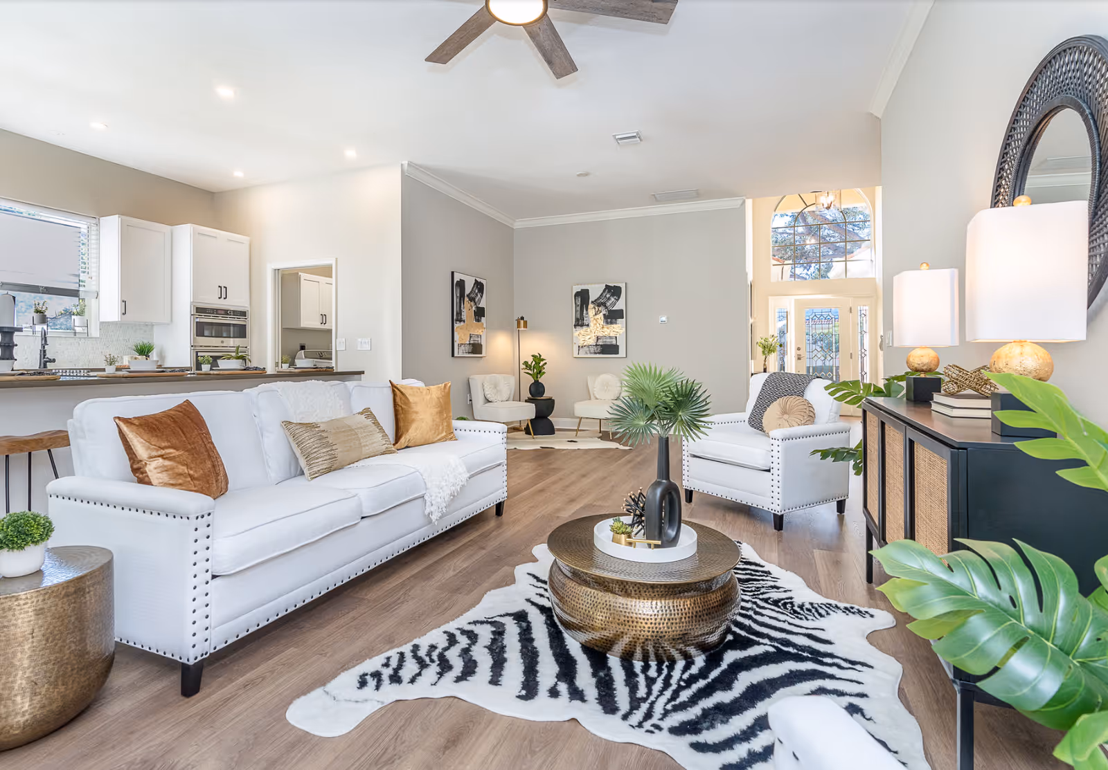 Bright and spacious living room with white sofas adorned with brown and beige cushions, a round metallic coffee table on a zebra-patterned rug, wooden flooring, a ceiling fan, and a sideboard with lamps and plants. The room opens to a kitchen area with white cabinets and bar stools, and a front door with glass panels is visible in the background.