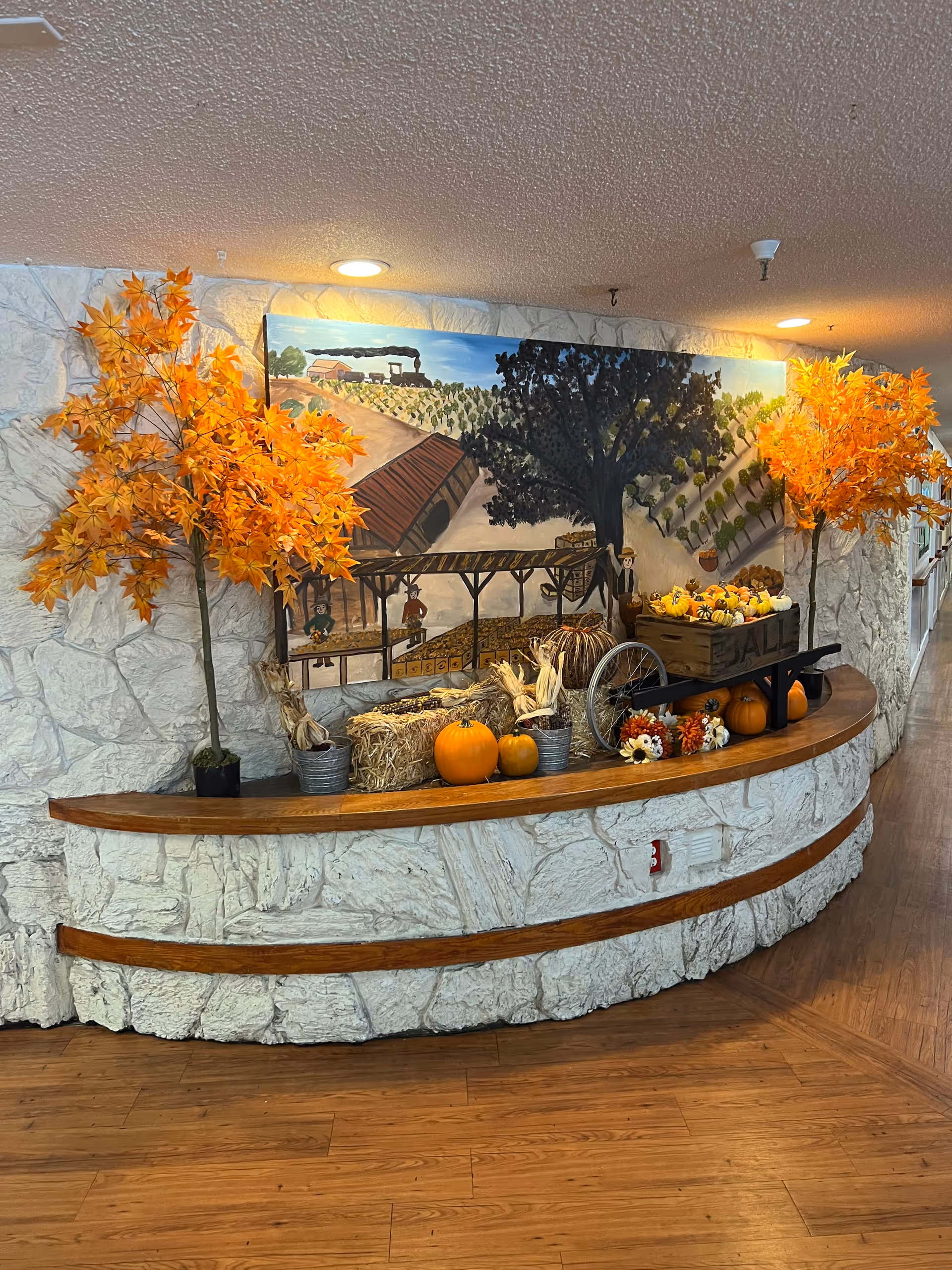 A curved stone wall with a wooden ledge decorated for fall with pumpkins, hay bales, artificial orange maple trees, and a wooden wheelbarrow filled with small pumpkins and gourds. Above the ledge is a large mural depicting a rural farm scene with a barn, trees, and a train in the background. The floor is wooden and the ceiling has recessed lighting.