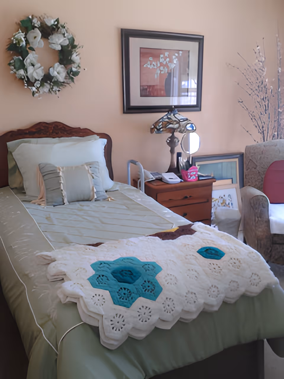 A neatly made single bed with light green bedding and decorative pillows in a cozy room. A crocheted blanket with blue and white patterns is draped over the foot of the bed. On the wall above the bed is a floral wreath and a framed picture. Next to the bed is a wooden nightstand with a decorative horse head lamp, a mirror, and some personal items. An upholstered armchair is partially visible on the right side of the image.