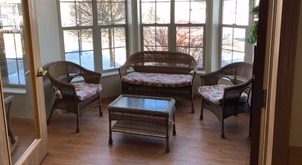 A cozy sunroom with large windows showing a snowy outdoor scene. The room contains a wicker loveseat and two wicker armchairs with floral cushions, and a wicker coffee table with a glass top on a wooden floor.