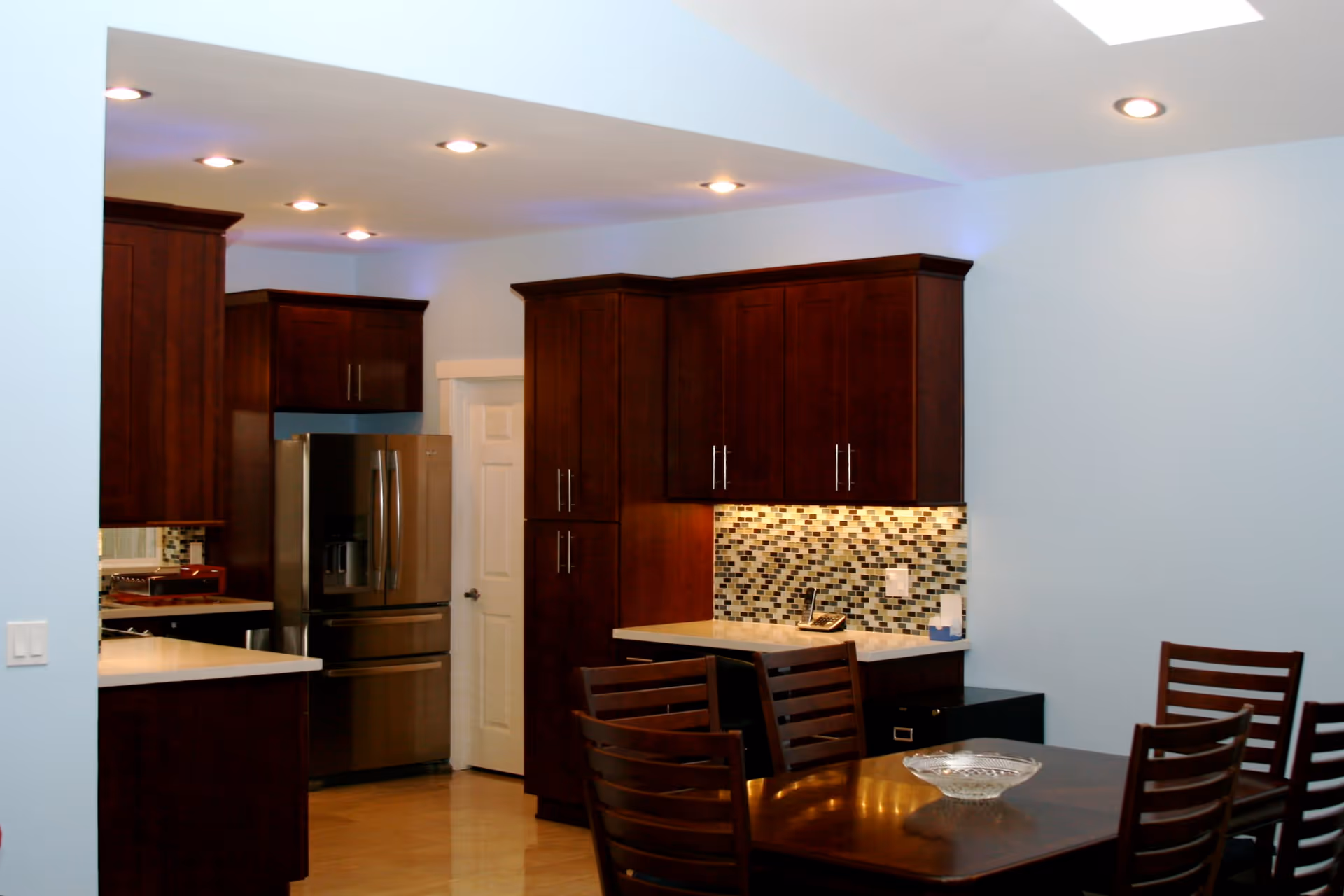 Interior view of a kitchen and dining area featuring dark wood cabinets, a stainless steel refrigerator, a mosaic tile backsplash, recessed ceiling lights, and a wooden dining table with chairs.