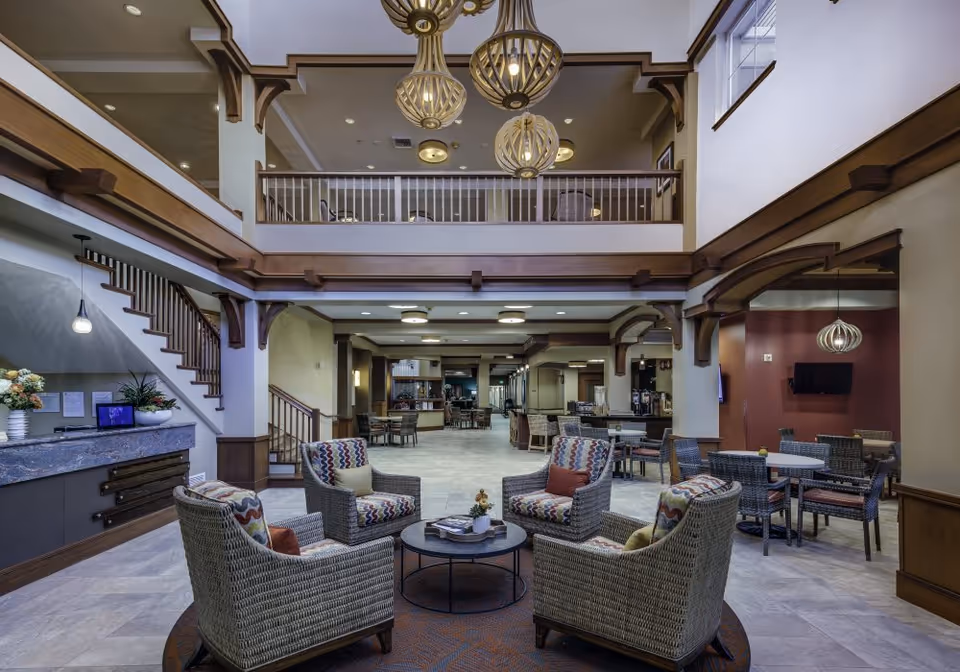 Spacious two-story senior living facility common area with wicker chairs around a coffee table, high ceilings, hanging chandeliers, and an upper balcony.