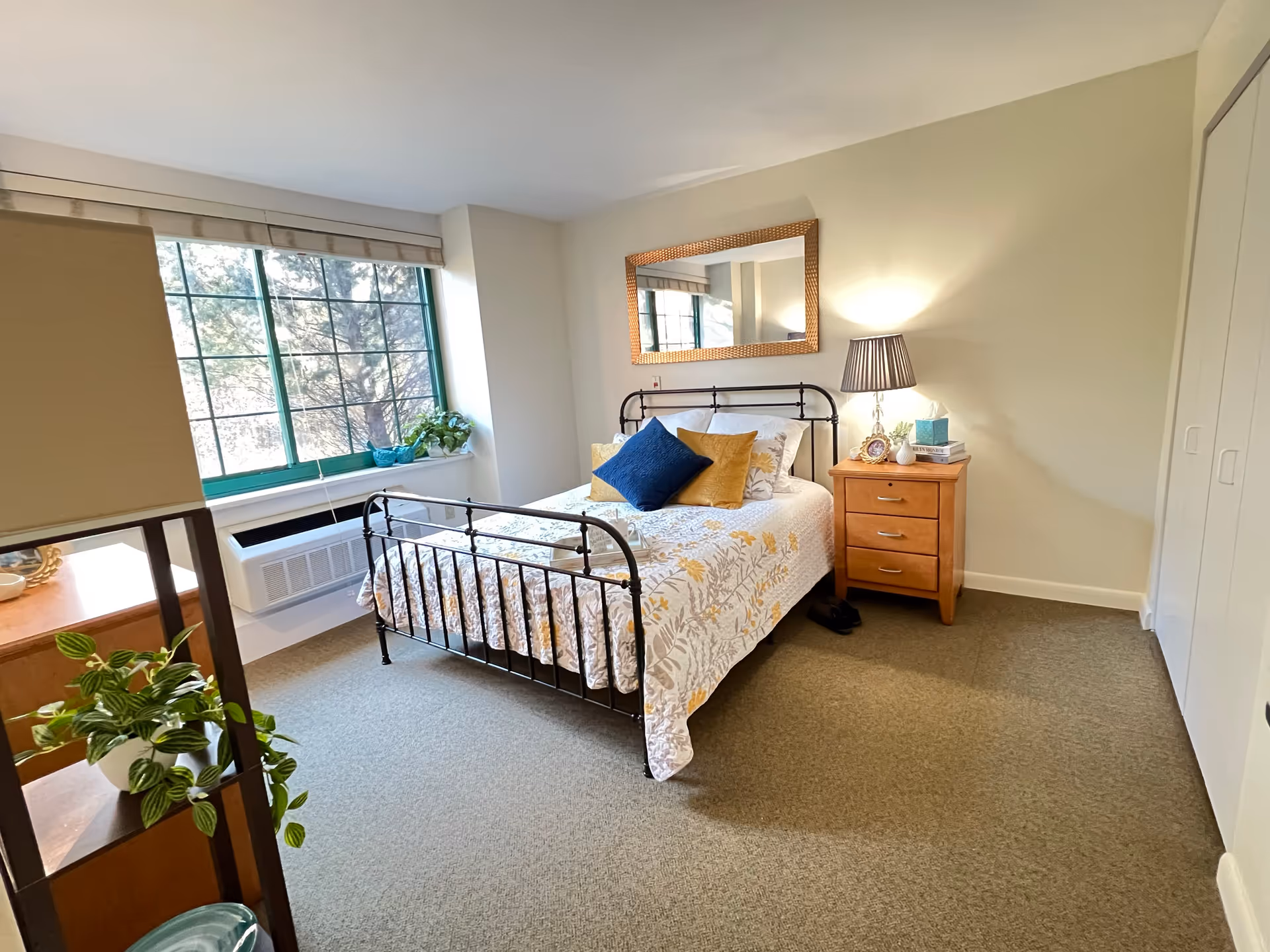 A bright bedroom with a metal-framed bed covered in a floral quilt and decorative pillows. There is a wooden nightstand with a lamp, books, and a photo frame next to the bed. A large window with green trim lets in natural light, and a mirror hangs on the wall above the bed. A shelving unit with plants is partially visible in the foreground.