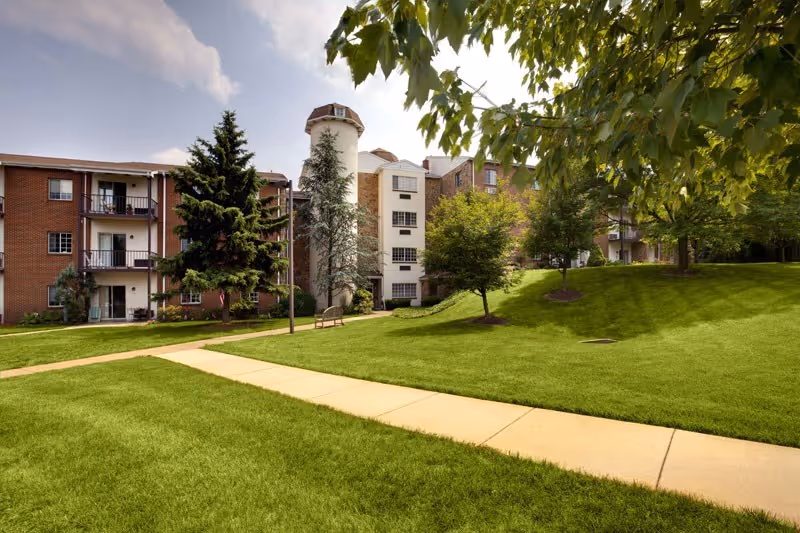 Well-manicured lawn and walkway leading to a multi-story brick senior living building with balconies and trees.