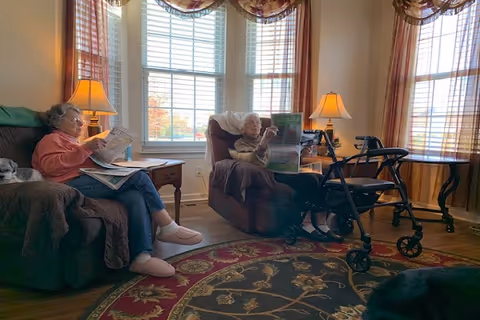 Two elderly women sitting in a cozy living room with large windows and curtains. One woman is seated on a couch reading a newspaper, and the other is in a recliner chair also reading a newspaper. There are two table lamps on side tables, a walker near the recliner, and a patterned area rug on the wooden floor.