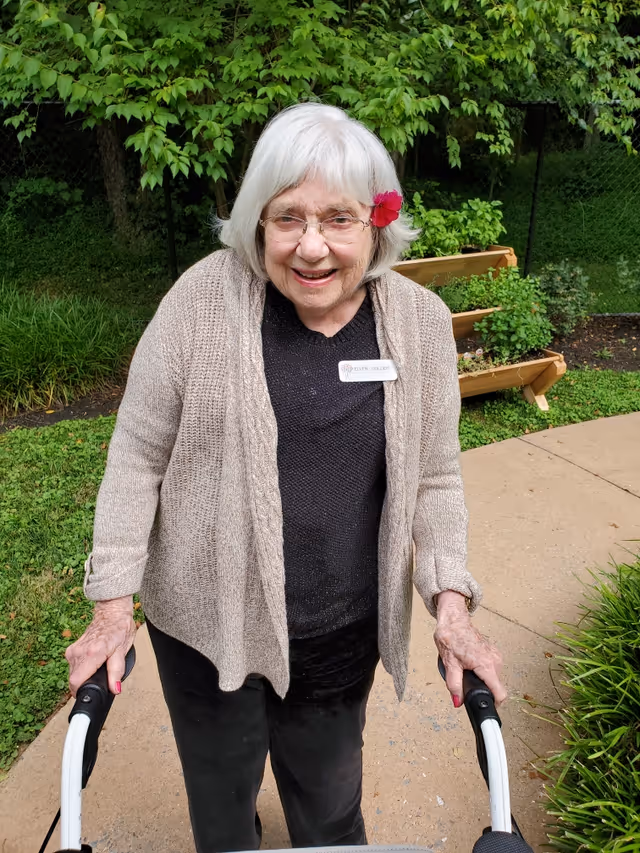 An elderly woman with white hair and glasses, wearing a beige cardigan and black top, smiling while standing outdoors on a paved path with greenery and plants in the background. She is holding onto a walker and has a pink flower tucked behind her ear.