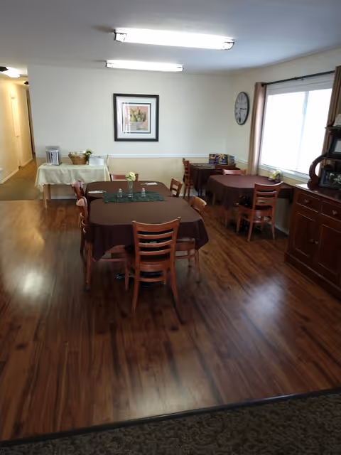 A dining area with wooden floors and multiple tables covered with brown tablecloths. Each table is surrounded by wooden chairs. There is a side table with a basket and a coffee maker against the wall, a framed picture hanging above it, and a large window with curtains letting in natural light. A clock is mounted on the wall near the window.