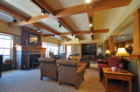 A spacious living room area in Windsor Manor featuring patterned armchairs, a sofa, wooden beams on the ceiling, a fireplace with a mirror above it, large windows letting in natural light, and several lamps providing warm lighting.