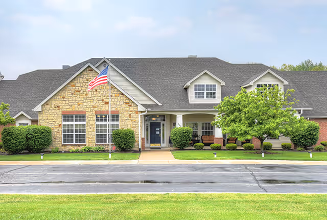Front exterior of a single-story senior living building with a stone and siding facade, American flag, landscaping, and a covered entrance.