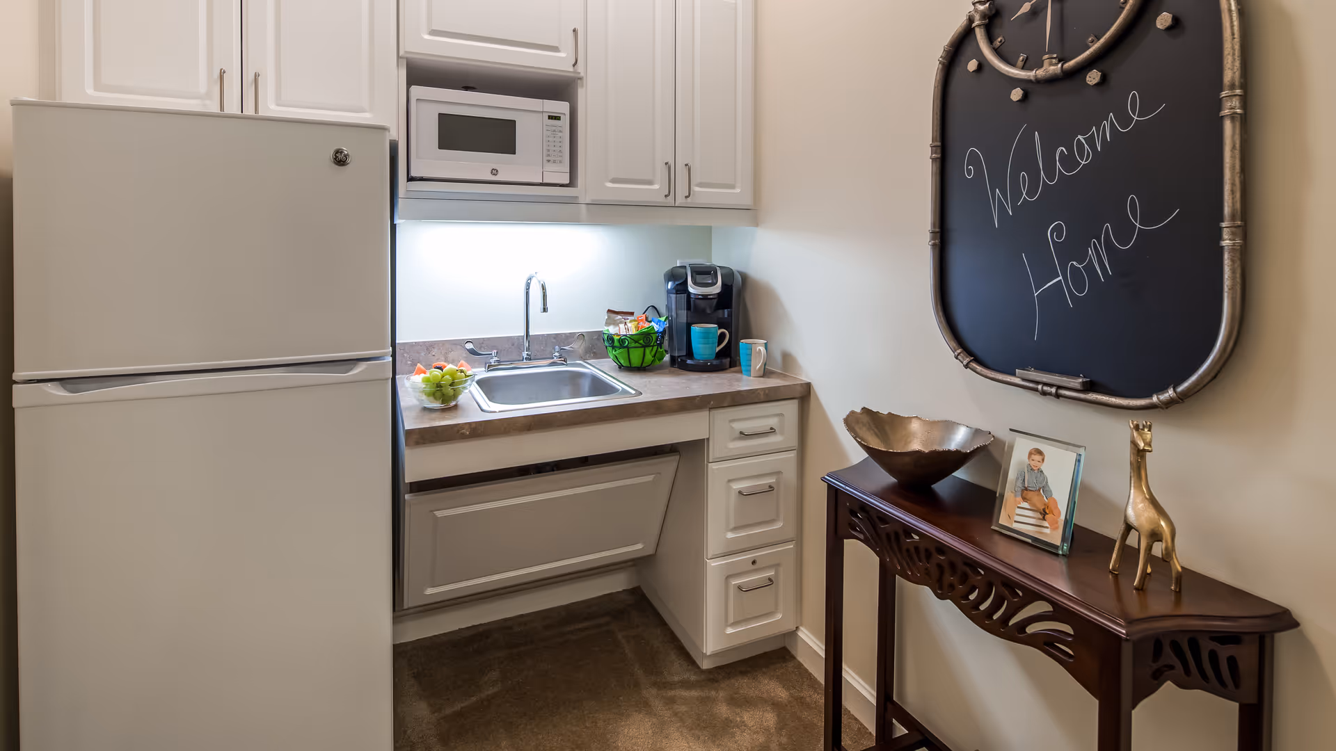 A small kitchen area with white cabinets, a refrigerator, a microwave, a sink, and a coffee maker on the countertop. To the right, there is a decorative wooden table with a metal bowl, a framed photo of a child, and a small giraffe figurine. Above the table, a chalkboard with the words 'Welcome Home' written on it is hanging on the wall.