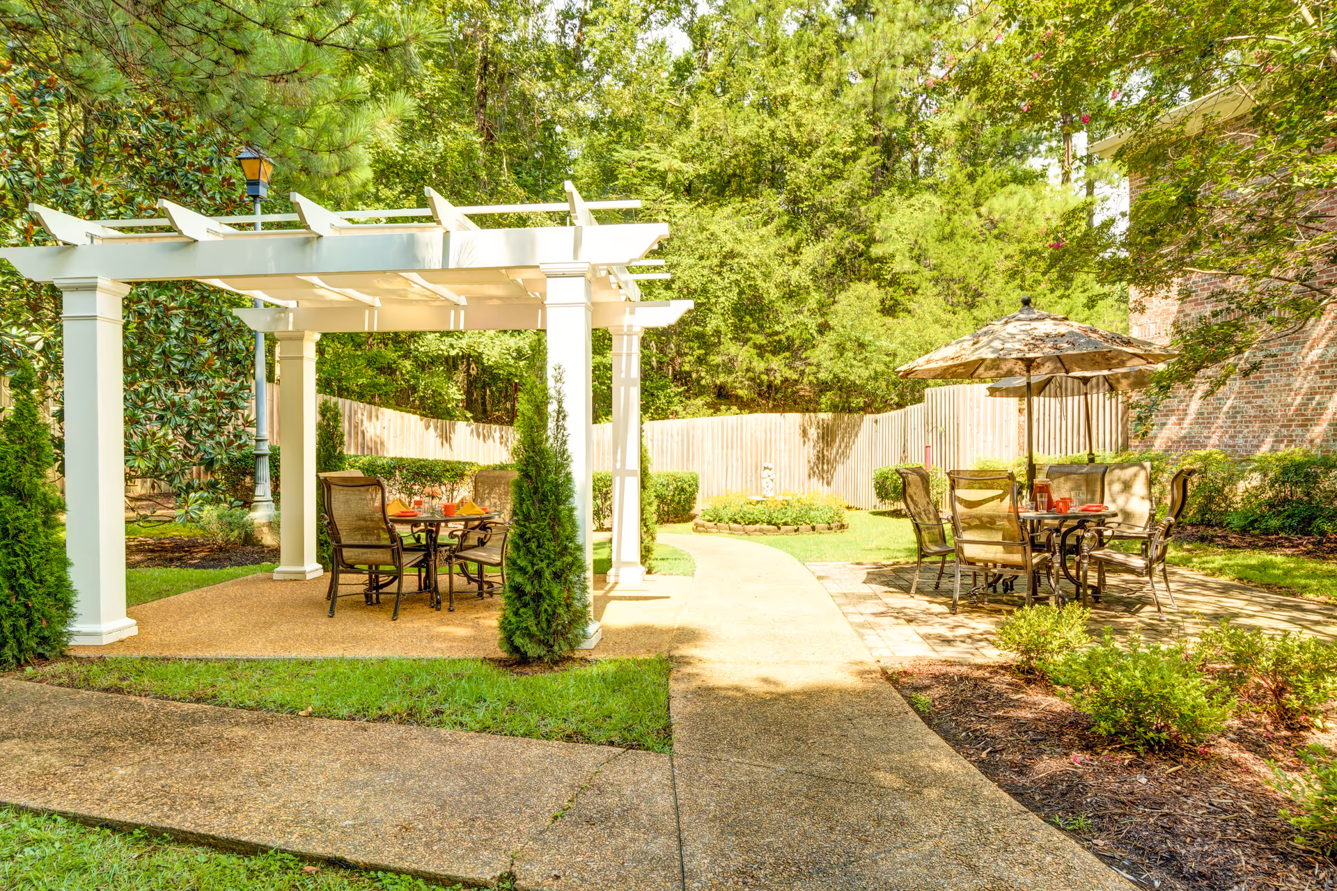Outdoor patio area with two seating sections; one under a white pergola with a table and chairs, and another with a table, chairs, and a large umbrella. The area is surrounded by green trees, bushes, and a wooden fence.