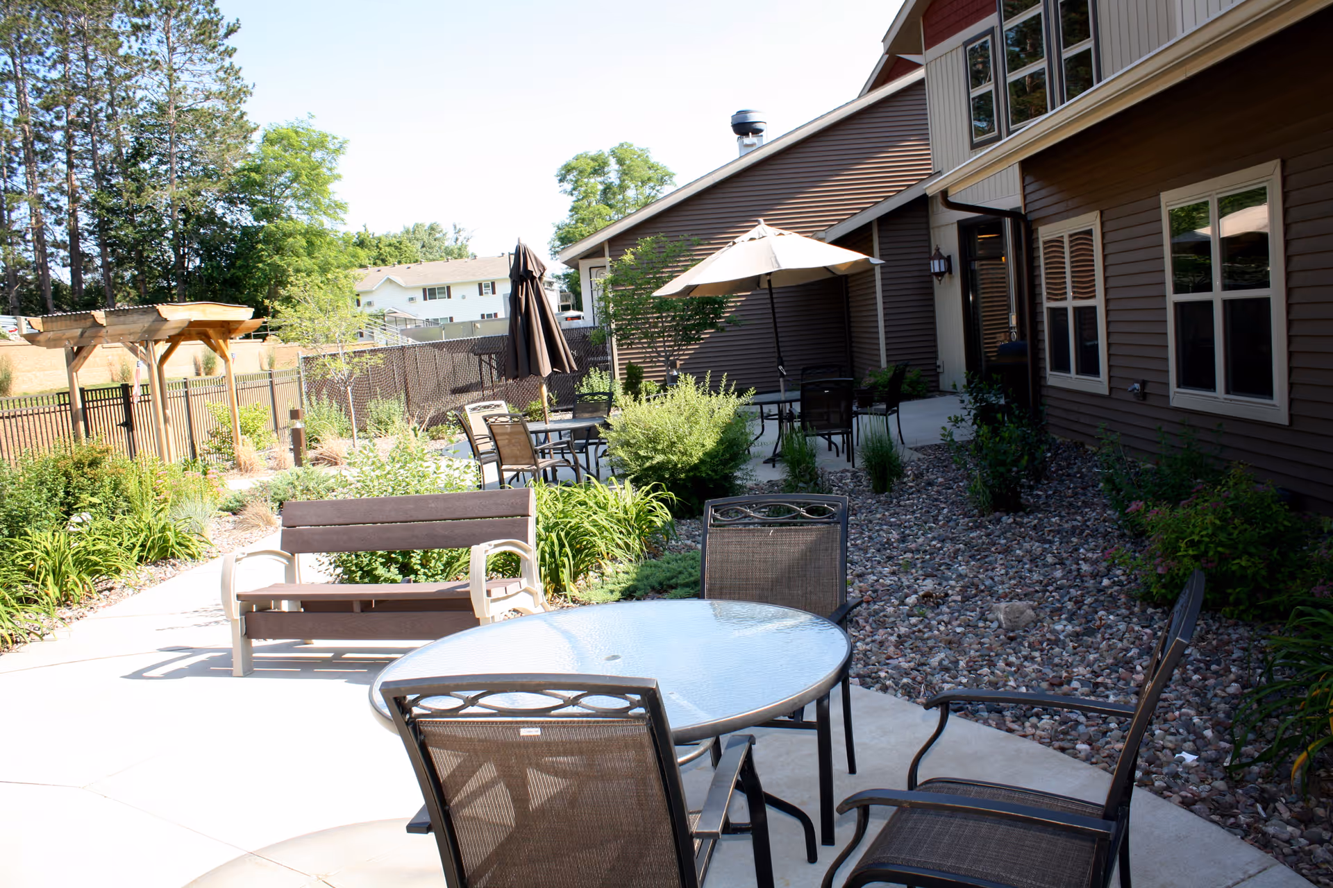 Outdoor patio area at Dove Healthcare – South Assisted Living with several metal and glass tables and chairs, a bench, umbrellas providing shade, surrounded by landscaping with bushes, rocks, and trees, adjacent to a brown building.