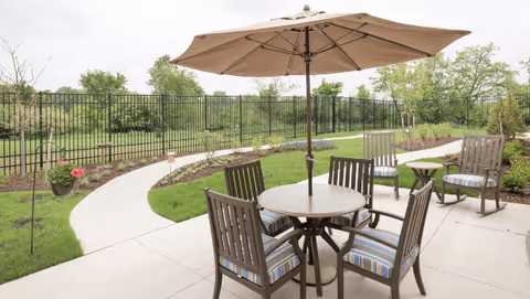 Outdoor patio area with a round table and four chairs with striped cushions under a large beige umbrella. Additional chairs and small tables are placed nearby on a concrete pathway surrounded by green grass, plants, and trees, enclosed by a black metal fence.