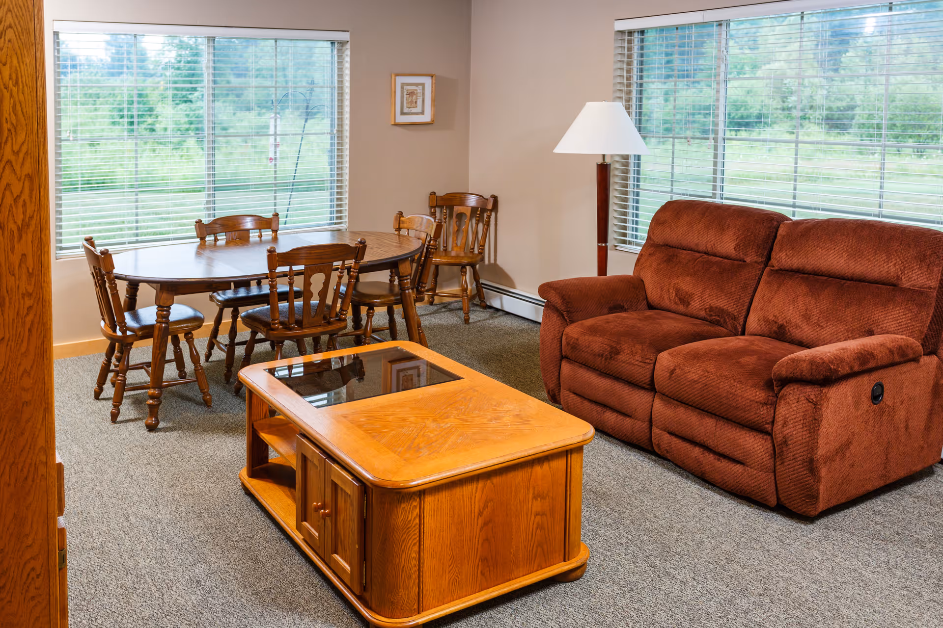 Light-filled living room with a brown upholstered reclining sofa, wooden coffee table and a dining table with chairs by large windows.