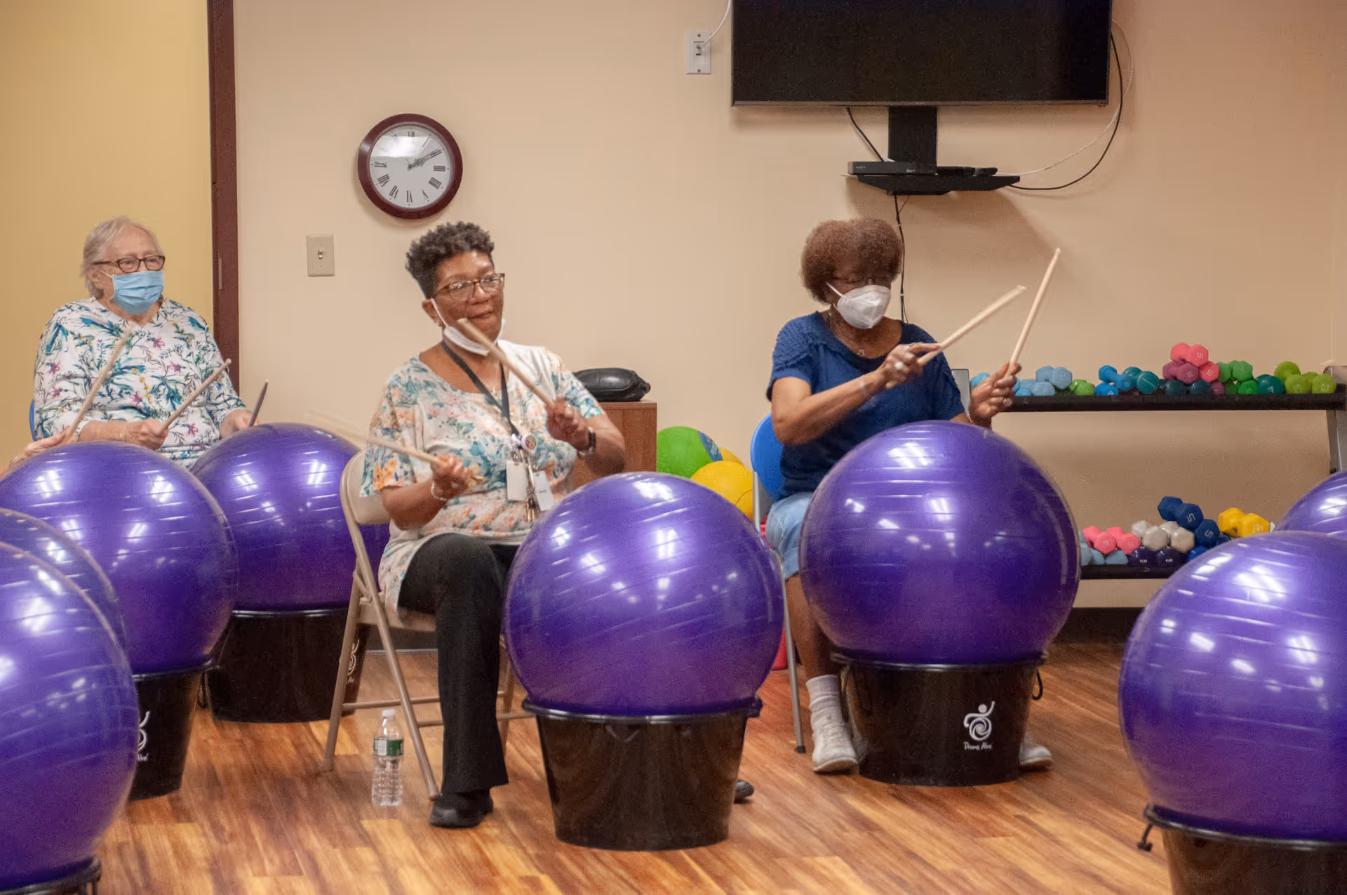 Three elderly women sitting on chairs in a room, each playing on large purple exercise balls with drumsticks. Two of the women are wearing face masks. Behind them is a table with colorful dumbbells and a wall-mounted TV.