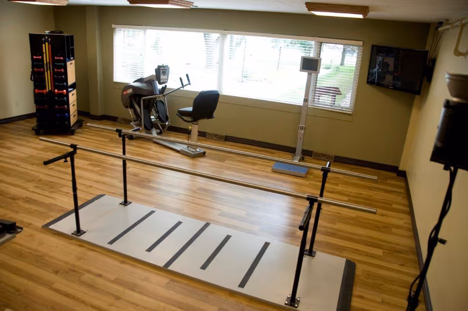 Bright exercise/therapy room with parallel bars, a recumbent bike, weight rack and large window.