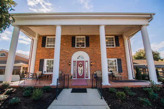 Front entrance of a two-story brick building with large white columns, a red door with a wreath, and porch seating.
