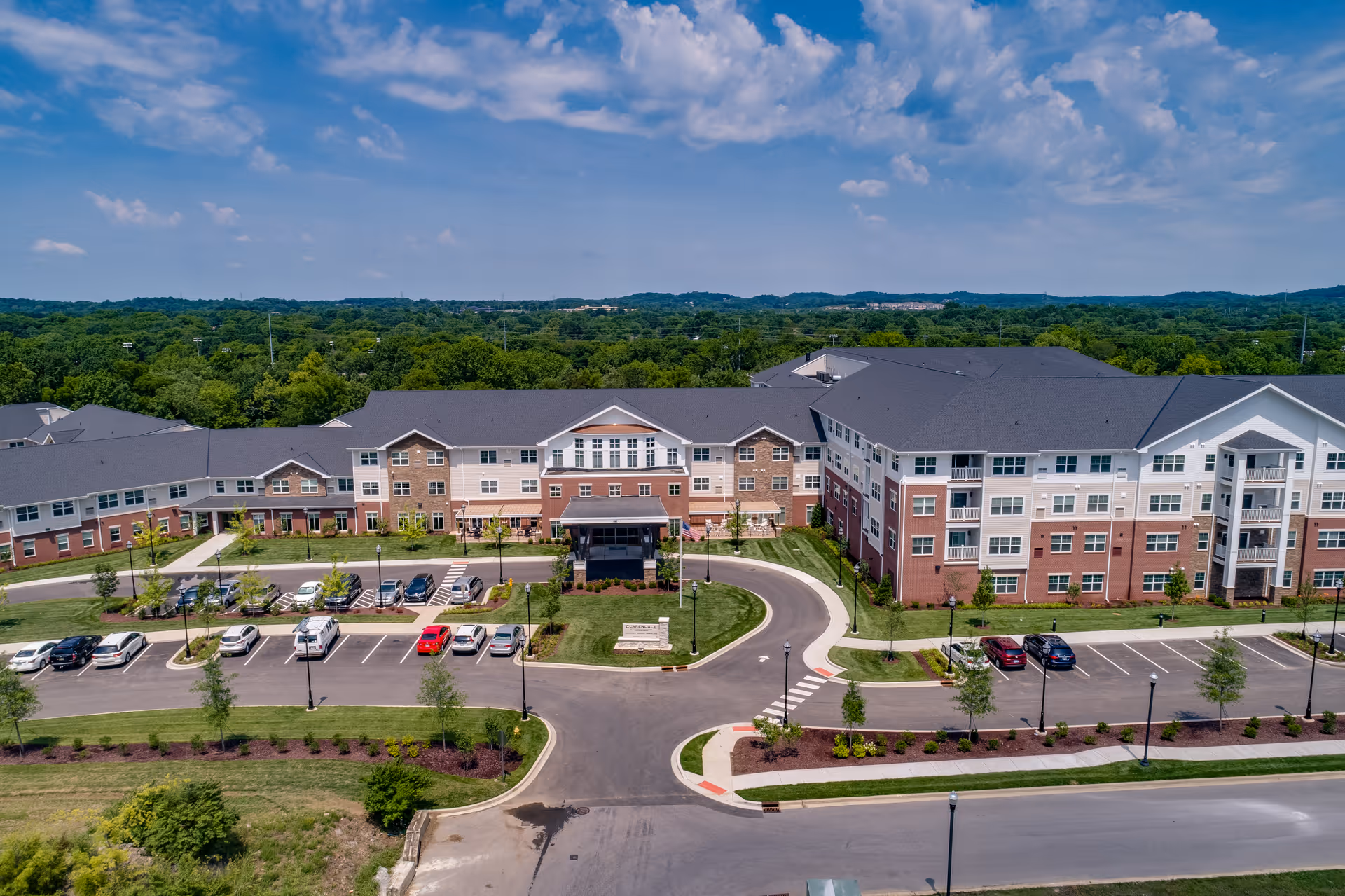 Aerial view of the Clarendale at Indian Lake senior living building with landscaped grounds, a circular drive and parking lot under a partly cloudy sky.