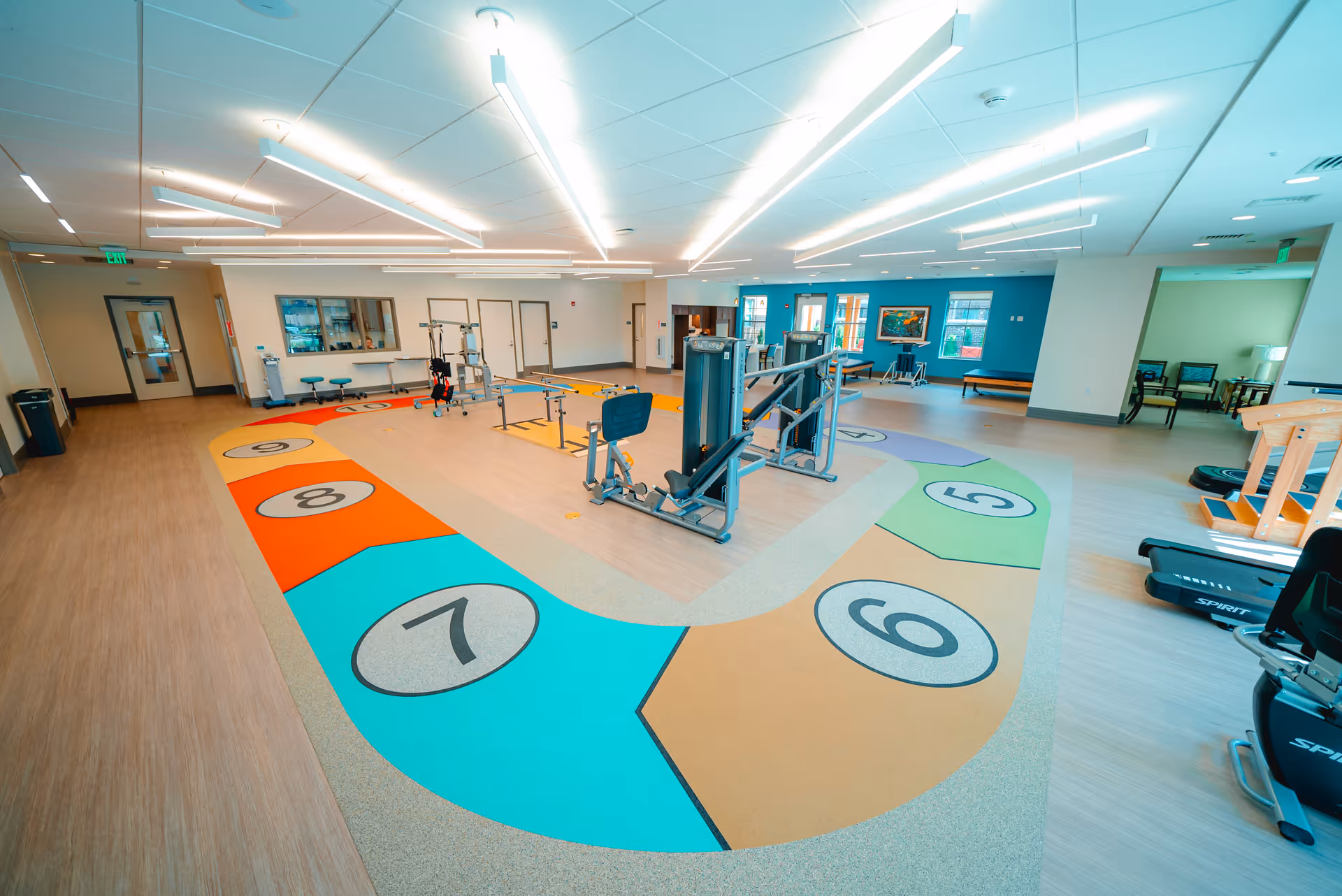Bright rehabilitation/exercise room with a colorful numbered floor track and various fitness machines and equipment.