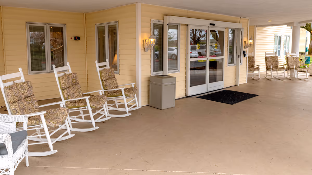 Covered entrance area of a senior living facility with several white rocking chairs featuring patterned cushions arranged along the yellow exterior walls. There are two wall-mounted lantern-style lights, a trash bin, and glass automatic sliding doors leading inside. Cars and trees are visible through the glass doors.