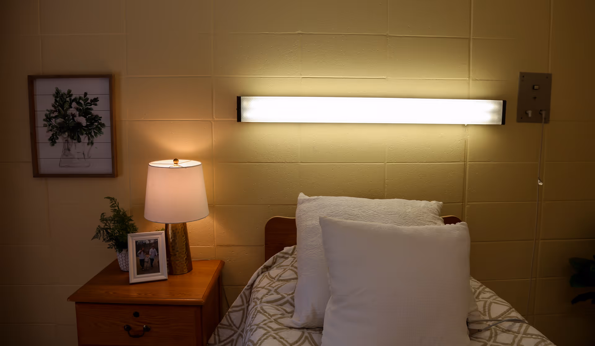 A neatly made single bed with white pillows under a wall-mounted light beside a wooden nightstand holding a lamp, framed photo and plant.