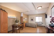 A well-lit senior living facility room with a single bed against the wall, a wooden table with two chairs, a tall wooden cabinet, a window with blinds, and decorative flowers on a small table. The room has light-colored walls and wood flooring.