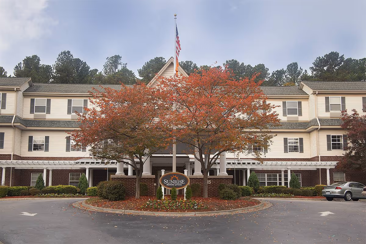 Exterior front view of a multi-story senior living facility named Sunrise at North Hills, featuring a circular driveway with landscaped trees showing autumn foliage and an American flag on a flagpole in front of the building.