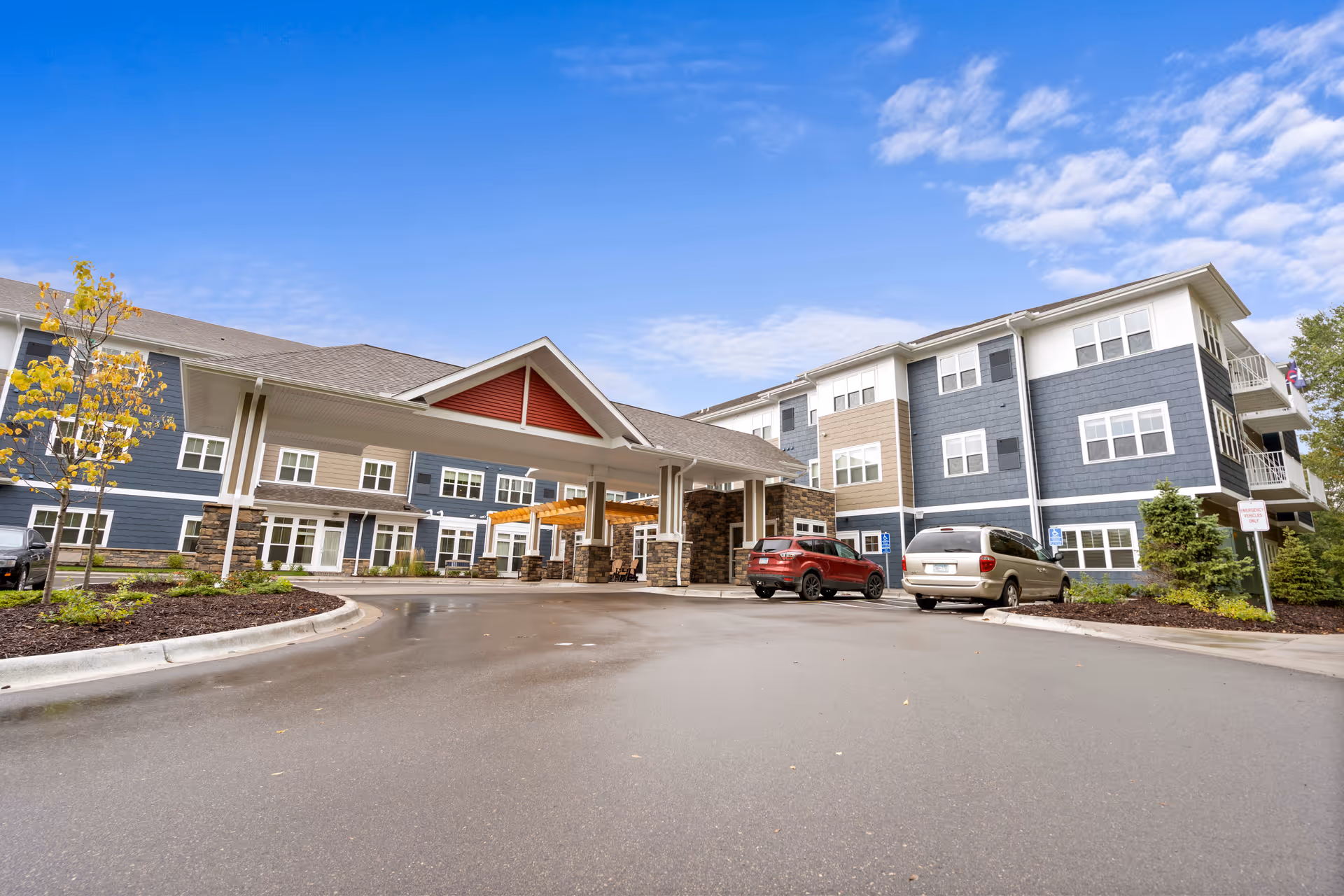 Exterior view of a modern senior living facility with a covered entrance, multiple windows, and a few parked cars in front. The building has blue and beige siding with stone accents and a clear blue sky above.