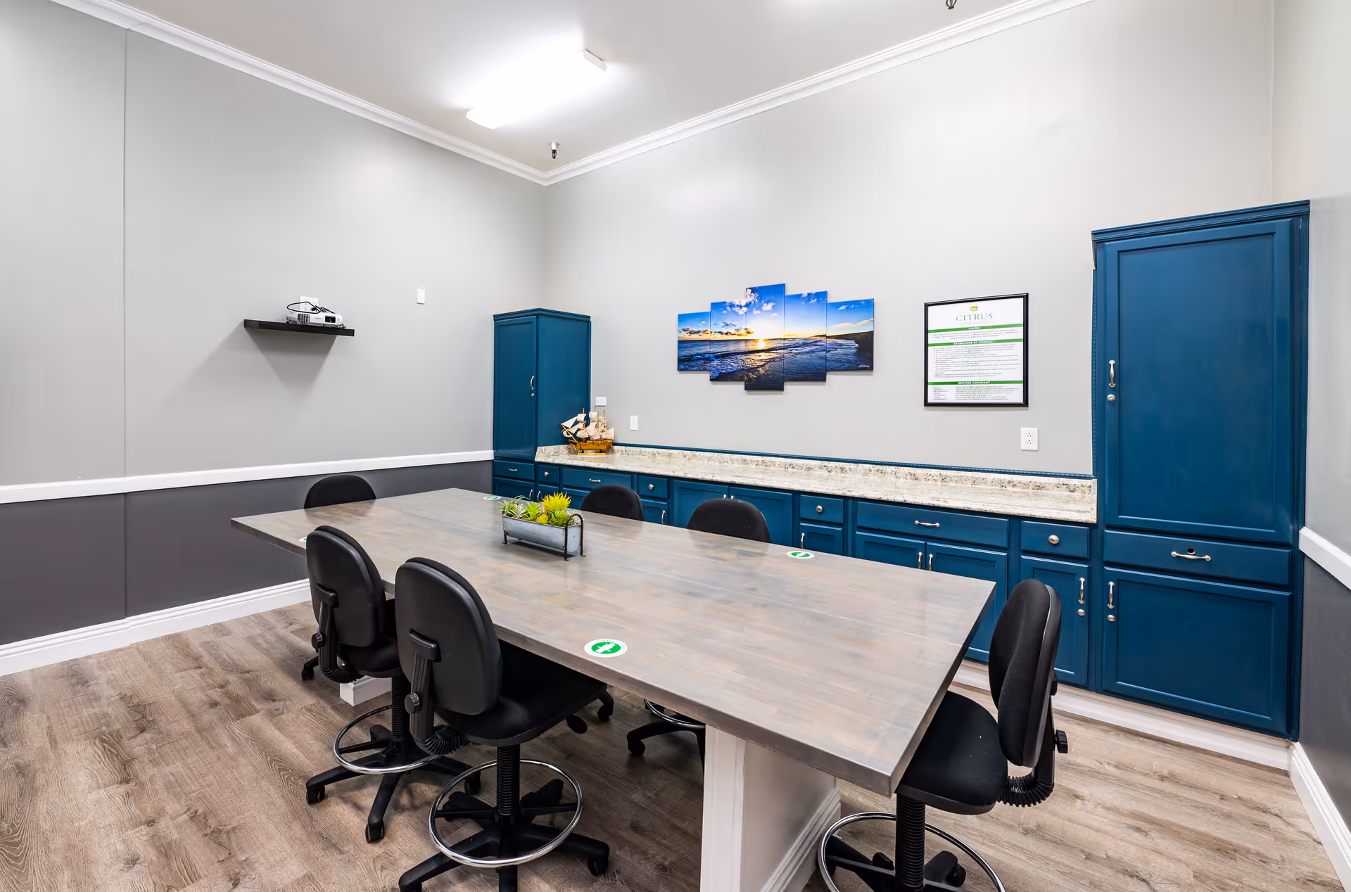 A meeting or conference room with a large rectangular wooden table surrounded by six black office chairs on wheels. The room has blue cabinets along one wall with a granite countertop, a multi-panel wall art depicting a sunset over the ocean, and a framed document on the wall. The floor is wood-patterned, and the walls are painted gray with white trim.