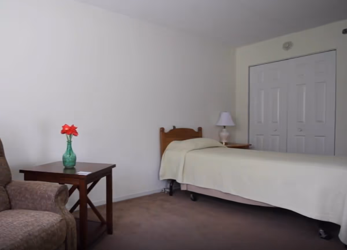 A simple bedroom with a single bed covered in a light beige blanket, a wooden headboard, a nightstand with a white lamp, a wooden side table with a green vase holding red flowers, and a brown upholstered armchair. The room has white walls and a closed closet with double doors.