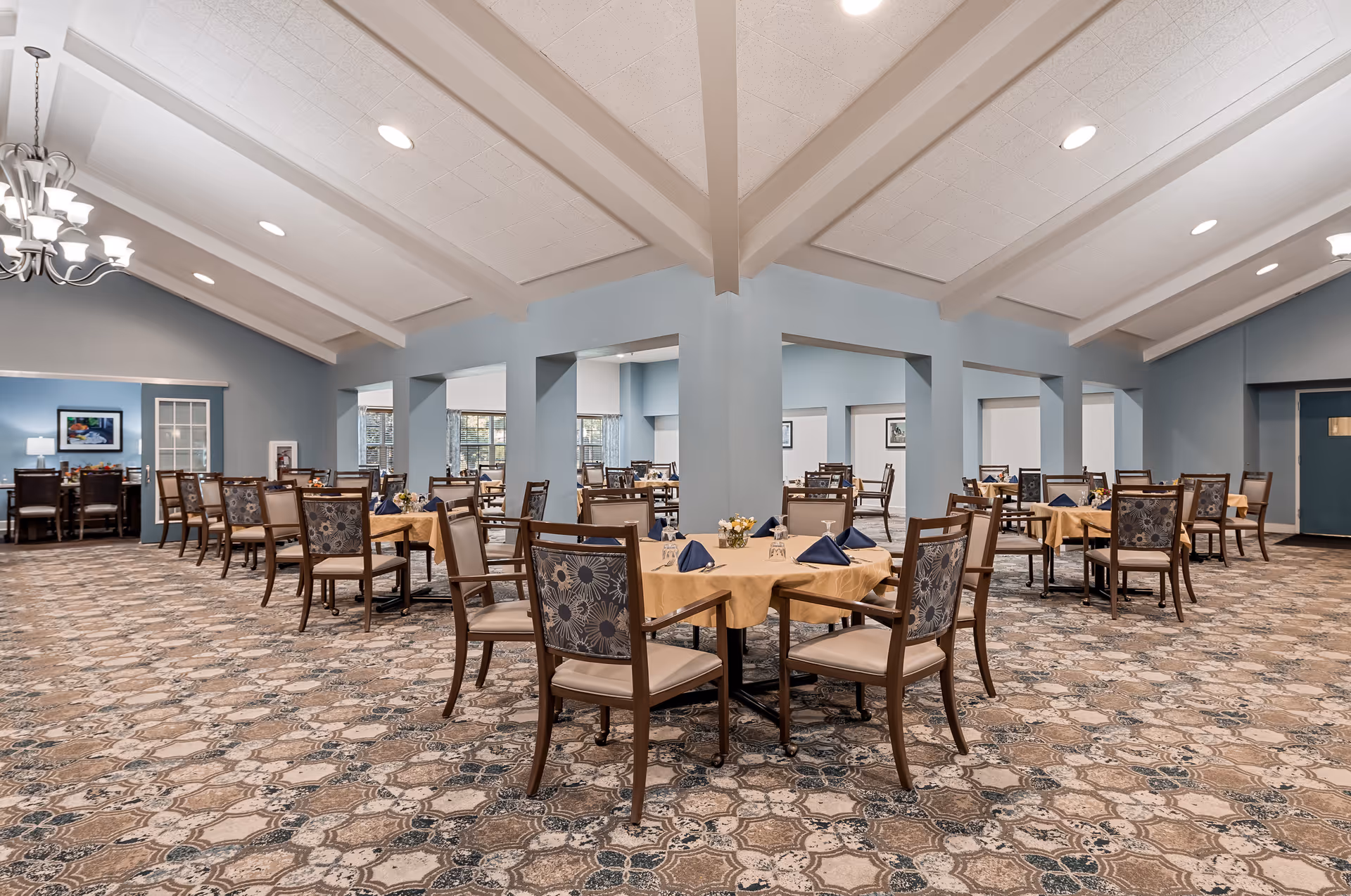 Spacious dining room with multiple tables covered in beige tablecloths and set with blue napkins. The room features patterned carpet, light blue walls, white ceiling beams, and a chandelier. Several chairs with floral upholstery surround the tables, and windows allow natural light to enter.