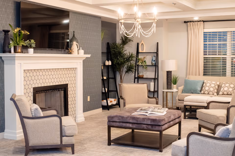 A cozy living room with a white fireplace featuring a patterned tile surround and a large flat-screen TV mounted above it. The room has beige upholstered armchairs and a sofa with decorative pillows, a large square ottoman with an open book on top, a side table with a lamp and plant, and two black ladder-style shelves with decorative items. A chandelier hangs from the ceiling, and a window with beige curtains is visible in the background.
