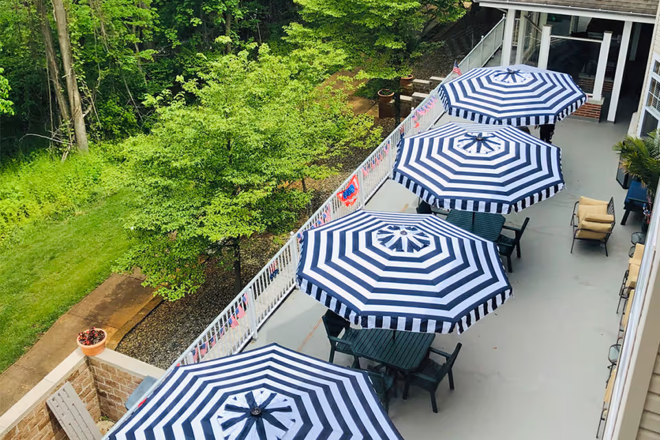 Outdoor patio area with four tables, each shaded by a blue and white striped umbrella. The patio is adjacent to a building and overlooks a green grassy area with trees. There are chairs around the tables and some additional seating along the building wall.