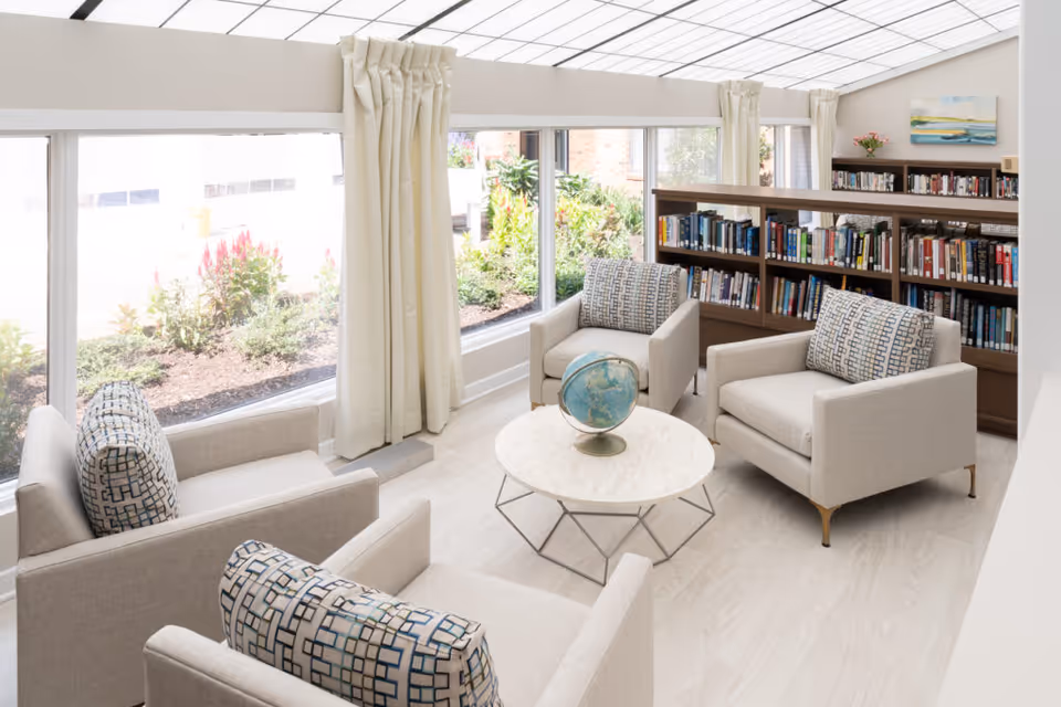 A bright and cozy sitting area in a retirement community with four beige armchairs arranged around a round white coffee table that holds a globe. Behind the chairs is a wooden bookshelf filled with books, and large windows with cream-colored curtains let in natural light and offer a view of a garden outside.