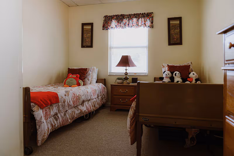 A small bedroom with two single beds, each with patterned bedspreads and stuffed animals on top. A wooden nightstand with a lamp is positioned between the beds under a window with a floral valance. Two framed pictures hang on the wall beside the window.