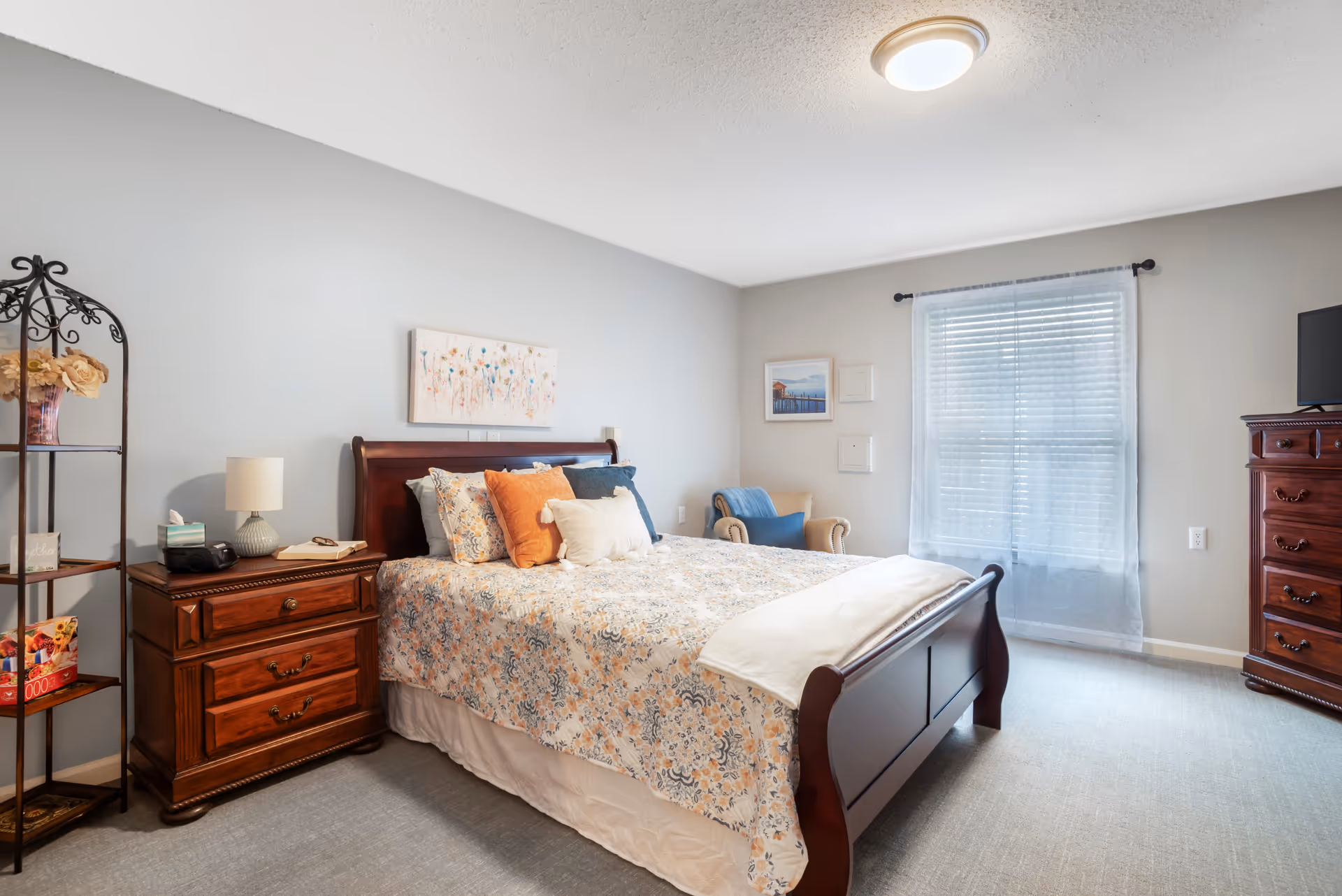 A well-lit bedroom with a wooden bed frame and floral patterned bedding with multiple pillows. There is a wooden nightstand with a lamp, tissue box, and a book beside the bed. A metal shelf with decorative items is on the left side. A window with sheer curtains lets in natural light, and a cushioned armchair with a blue throw is placed near the window. A wooden dresser with a TV on top is on the right side of the room. The walls are painted light gray and decorated with framed pictures.