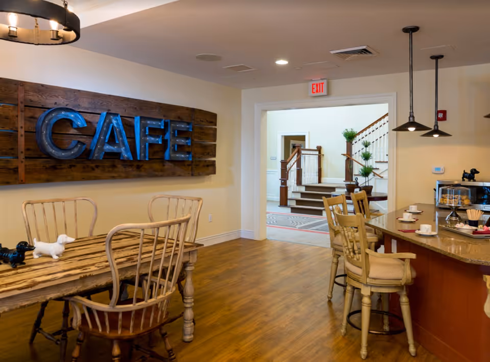 Interior café area with wooden tables and chairs, a large 'CAFE' sign on the wall, and a counter with barstools near an open doorway to a staircase.