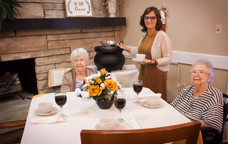 Two elderly women sitting at a dining table set with cups, saucers, and glasses of dark beverage, with a woman standing behind them serving soup from a black pot. The room has a stone fireplace with a sign above it that reads 'Be Our Guest' and a floral centerpiece on the table.