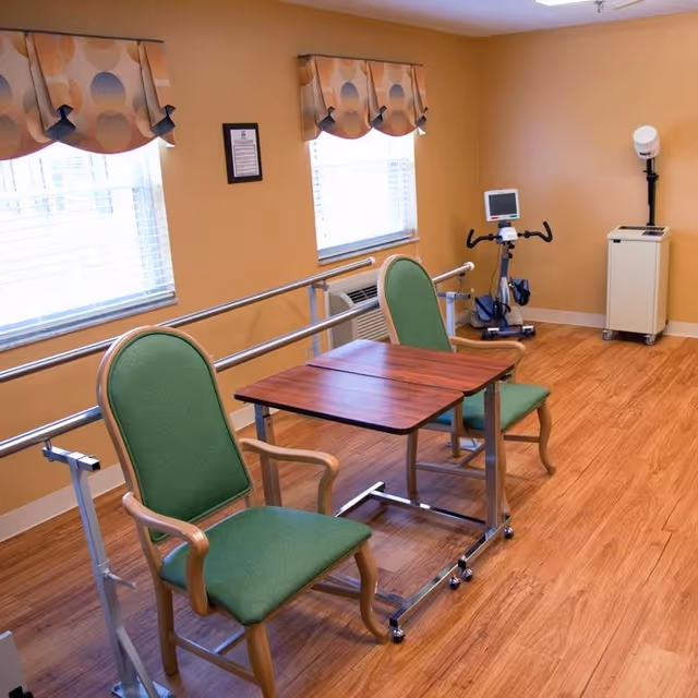 A rehabilitation room with two green cushioned chairs and a wooden table on wheels between them. The room has wooden flooring, two windows with patterned valances, handrails along the wall, and exercise equipment including a stationary bike and a medical device on a stand.