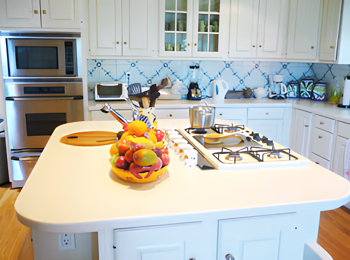 A bright kitchen with white cabinets and a large island countertop. The island features a built-in gas stove and a colorful fruit basket with various fruits. The kitchen also includes a double oven, a microwave, a blender, a kettle, and other small appliances. The backsplash has a blue and white tile pattern.