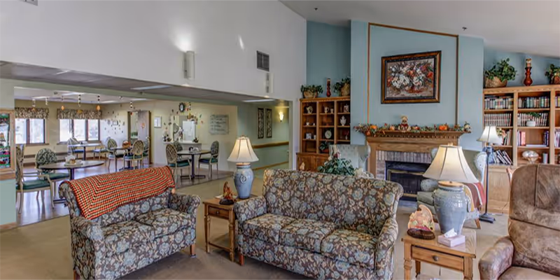 Common living room with patterned sofas, side tables and lamps facing a fireplace and bookshelves, with a dining area visible in the background.
