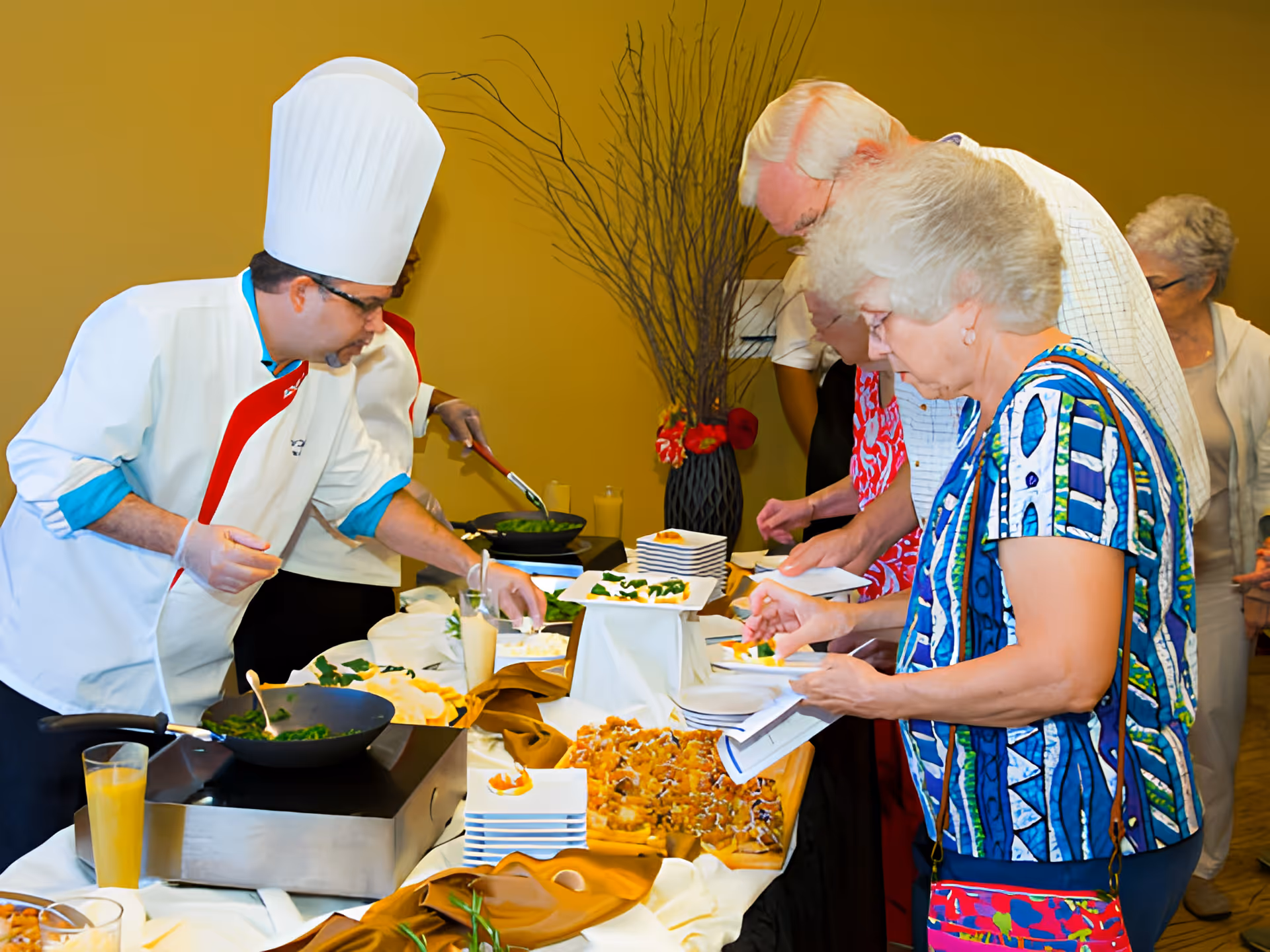 A chef wearing a white uniform and tall white chef hat is serving food to a group of elderly people at a buffet table. The table is covered with a white and gold tablecloth and has various dishes and plates arranged on it. The elderly individuals are selecting food and holding plates, with a decorative vase with branches and flowers in the background.