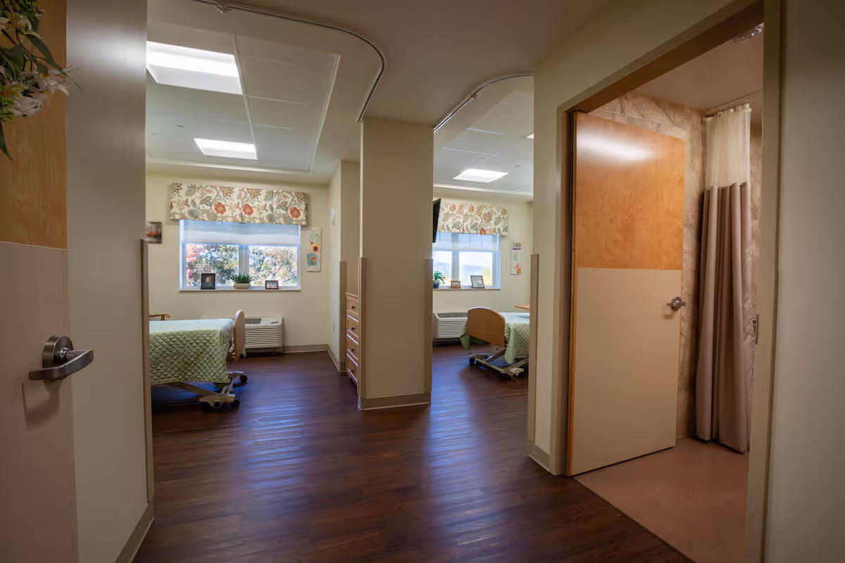 Interior view of a nursing facility room with two hospital beds, each with green bedspreads, positioned near windows with floral valances. The room has wooden flooring and neutral-colored walls. To the right, there is an open door leading to a bathroom with a shower curtain visible.